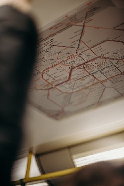A close-up photograph of a detailed tube map of London, featuring numerous coloured lines and stations, held by a person during a home relocation or moving planning session. The map is inside a booklet or folded sheet, with the person's fingers gripping the edges while they study the routes. The surrounding environment includes a slightly blurred ceiling light and the inside of a vehicle or a room, suggesting a transport context. The image emphasizes careful planning of a furniture transport or house removals process, which is relevant to the services offered by Man with Van Earlsfield for seamless moving logistics and packing and moving tasks.