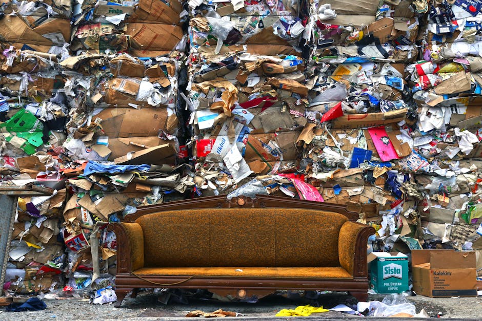 A vintage brown upholstered sofa with wooden trim is placed on a pavement in front of a large pile of mixed household waste and recyclable materials, including cardboard boxes, paper, plastic packaging, and fabric remnants. The waste is stacked against a wall and covers the entire background of the image. Nearby, there are additional cardboard boxes labeled 'Sista' and 'Kalekim.' The scene is outdoors with natural lighting and no visible moving equipment. This setup may relate to a home relocation or waste clearance process, as it depicts the staging area for removal services with Man with Van Earlsfield involved in loading or disposal activities.
