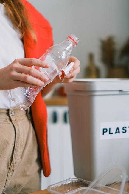 A person wearing a white top and beige trousers is standing next to a grey plastic recycling bin labeled 'PLASTIC' in a room with neutral walls. They are holding a clear plastic bottle with a red cap, which appears to contain a small amount of liquid, and are in the process of discarding it into the bin. The scene is well-lit with natural or artificial light, emphasizing the act of waste disposal. The image reflects environmentally conscious habits related to waste separation and recycling, aligning with discussions about home organization or packing during a move. In the background, there may be additional household items, but they are out of focus, keeping the emphasis on the person and the recycling activity.