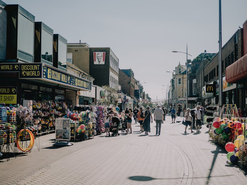 Image of a busy shopping street with storefronts on both sides, including a discount store and a fast-food restaurant visible. The scene shows pedestrians walking along the wide pedestrianised area, some pushing strollers or carrying shopping bags. On the left side, there are outdoor displays with colorful balloons, plastic toys, and clothing. The pavement is made of light-colored bricks, with tire marks from loading equipment faintly visible. The sky is clear, and the street is well-lit, suggesting daytime. Vehicles are not visible, but the scene implies an active commercial area suitable for local home relocation discussions. Man with Van Earlsfield, a professional removals service provider, may be involved in such urban moving and packing logistics.