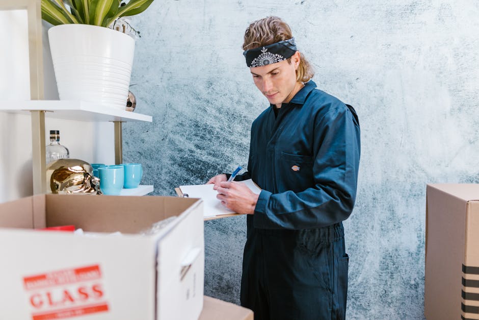 A professional man with dark skin and an afro hairstyle, wearing a blue t-shirt with a company logo, is taking notes on a clipboard while standing in an indoor space surrounded by packing materials. S