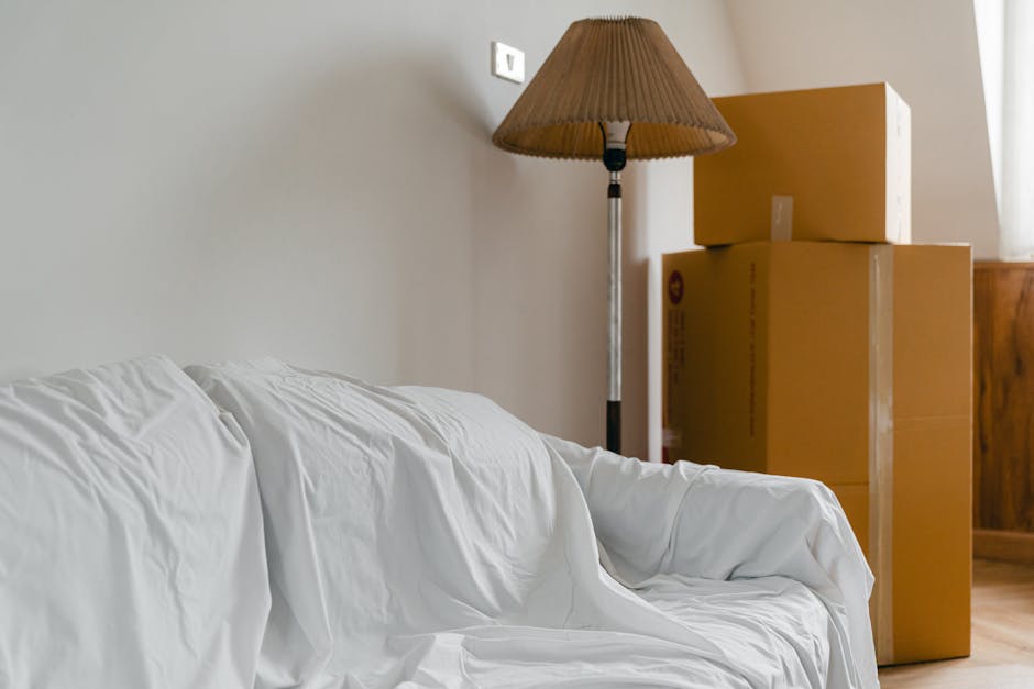 Inside a residential room, several packed cardboard boxes of varying sizes are stacked near a wooden cabinet, positioned close to a wall. A tall standing lamp with a beige pleated fabric shade is situated next to the boxes, casting a subtle light on the scene. In the foreground, a white sofa draped with a slightly wrinkled white sheet is visible, indicating an ongoing house removal or packing process. The scene captures part of the interior environment with natural lighting coming from a window out of view, highlighting the preparation stage of a home relocation. The presence of packed boxes, protective wrapping, and the arrangement suggest furniture transport and unpacking activities associated with professional removals, such as those offered by Man with Van Earlsfield, experienced in packing and moving services, including the loading process involved in house moves.