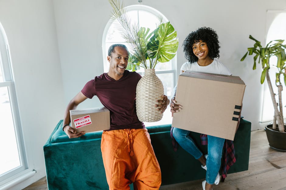 A woman with long, curly, light brown hair is writing the word 'FRAGILE' on a large cardboard moving box using a black marker. The box is situated on top of several other packed boxes inside a room wi