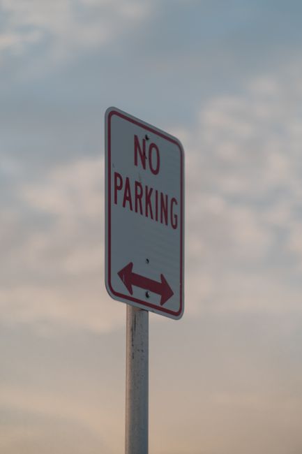A tall, rectangular 'No Parking' sign with a white background and red border, mounted on a metal pole, situated on a paved street near a residential property in Earlsfield (SW18). The sign features red lettering reading 'NO PARKING' and has red arrow symbols pointing left and right beneath the text, indicating parking restrictions apply on both sides of the sign. In front of the sign, there is a white van with an open side door, positioned partially on the pavement and partially extending into the loading area outside a house. The van is being loaded or unloaded with cardboard boxes, plastic-wrapped furniture, and various packing materials, including padded blankets and plastic covers. Removal company staff, partially visible, are lifting or carrying some of these items towards the vehicle, using trolleys and straps as they carefully handle household belongings, indicating a home relocation or moving process. Bright, natural daylight illuminates the scene with a partly cloudy sky overhead, emphasizing the practical aspect of furniture transport and packing during a professional move by Man with Van Earlsfield.
