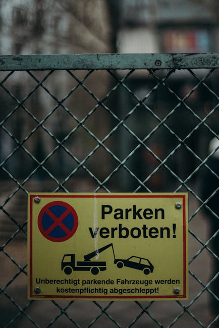 A tall, rectangular 'No Parking' sign with a white background and red border, mounted on a metal pole, situated on a paved street near a residential property in Earlsfield (SW18). The sign features red lettering reading 'NO PARKING' and has red arrow symbols pointing left and right beneath the text, indicating parking restrictions apply on both sides of the sign. In front of the sign, there is a white van with an open side door, positioned partially on the pavement and partially extending into the loading area outside a house. The van is being loaded or unloaded with cardboard boxes, plastic-wrapped furniture, and various packing materials, including padded blankets and plastic covers. Removal company staff, partially visible, are lifting or carrying some of these items towards the vehicle, using trolleys and straps as they carefully handle household belongings, indicating a home relocation or moving process. Bright, natural daylight illuminates the scene with a partly cloudy sky overhead, emphasizing the practical aspect of furniture transport and packing during a professional move by Man with Van Earlsfield.