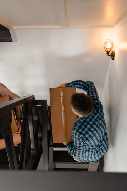 A man wearing a blue and black checkered shirt is inside a narrow staircase area of a flat in Earlsfield, carrying a medium-sized cardboard box. The staircase has black metal railings and wooden steps. The interior wall is painted white, with a decorative wall-mounted light fixture emitting warm light. The space appears to be part of a home relocation process, with the man positioned near a landing, preparing to move the box down the stairs. In the background, partially visible boxes and packing materials are situated on the stairs, indicating ongoing packing and moving preparations. The lighting is soft and warm, and the setting reflects a typical interior scene during furniture transport and removals, with [COMPANY_NAME] providing professional moving services for house relocations.