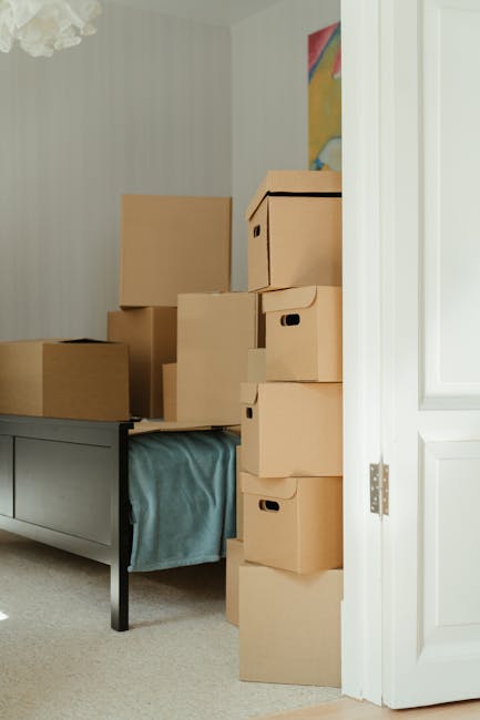 Interior of a room during a house relocation, featuring a stack of brown cardboard moving boxes of various sizes placed on a grey sideboard and on the floor next to the door. The boxes are packed with packing materials, some with black built-in handles, and are arranged in a stacked formation to optimize space. A light blue moving blanket or protective covering is partially visible beneath the boxes on the sideboard, indicating preparation for furniture transport. The room has a neutral colour palette with beige carpet flooring, light wallpaper, and a white door frame, which is slightly ajar, revealing part of a colourful abstract painting on the wall. The environment suggests an ongoing packing process as part of a home relocation or furniture moving service, illustrating the logistics of loading house items in a typical Earlsfield flat. The overall scene captures the careful placement and staging of items ready for transport, consistent with professional removals services by Man with Van Earlsfield.