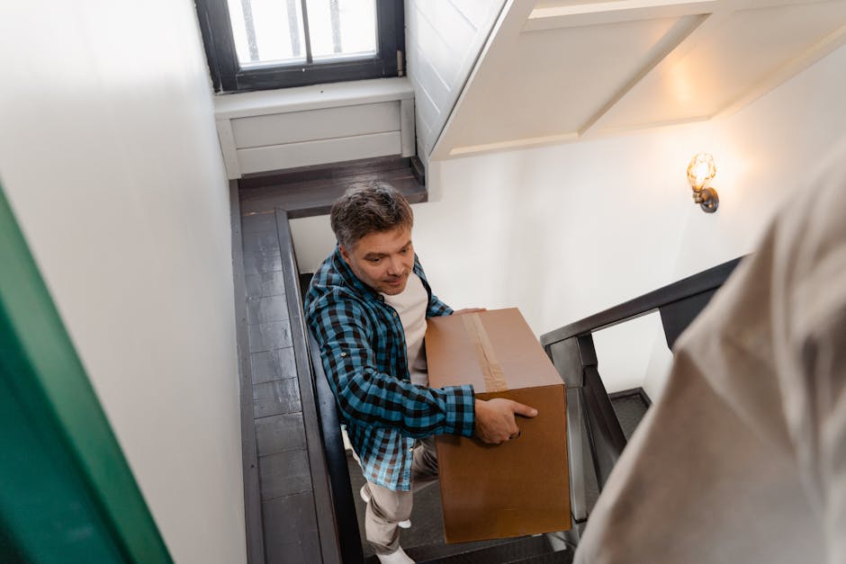A man wearing a blue and black checkered shirt is inside a narrow staircase area of a flat in Earlsfield, carrying a medium-sized cardboard box. The staircase has black metal railings and wooden steps. The interior wall is painted white, with a decorative wall-mounted light fixture emitting warm light. The space appears to be part of a home relocation process, with the man positioned near a landing, preparing to move the box down the stairs. In the background, partially visible boxes and packing materials are situated on the stairs, indicating ongoing packing and moving preparations. The lighting is soft and warm, and the setting reflects a typical interior scene during furniture transport and removals, with [COMPANY_NAME] providing professional moving services for house relocations.