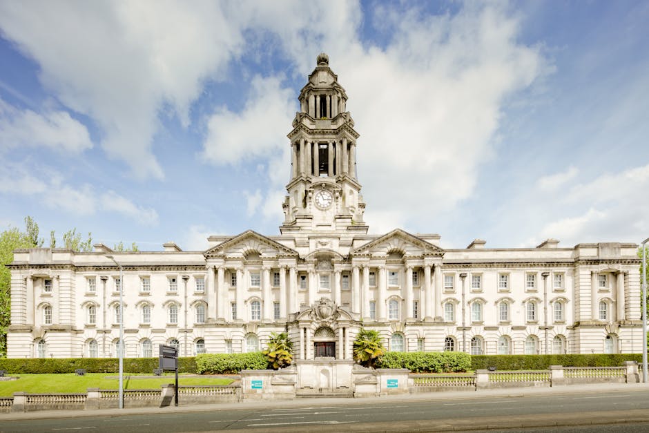 A large, historic white stone building with intricate architectural details, featuring a central clock tower topped with a small dome and decorative spires, situated along a tree-lined street under a partly cloudy sky. The building has multiple arched and rectangular windows, with the main entrance framed by columns and a decorative pediment. Surrounding the building is a well-maintained hedge and paved sidewalk. In front of the building, part of a grey van with its back doors open can be seen on the pavement, with visible packing materials such as cardboard boxes and plastic wrapping nearby, while a team of movers from Man with Van Earlsfield are loading household furniture, including wooden furniture and cushions, into the vehicle as part of a home relocation or furniture transport process. The scene exemplifies professional removal and moving logistics in an urban environment, consistent with house removals and packing services described on the Wandsworth Council permits page.