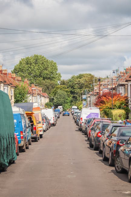 A residential street scene featuring a narrow asphalt road lined with closely parked cars on both sides, including various models in colors such as white, silver, blue, and black. The street is flanked by terraced houses with brick facades, some with small front gardens and hedges, and rooftops with chimneys. In the background, large green trees and hypothetically overcast skies with grey clouds are visible. Overhead, a network of power lines stretches across the sky. The image suggests a typical urban area suitable for house removals or home relocation services, as provided by Man with Van Earlsfield, with the loading process likely occurring in such neighbourhoods. The scene emphasizes the importance of careful planning when transporting furniture and boxes through tight residential streets during house moves.